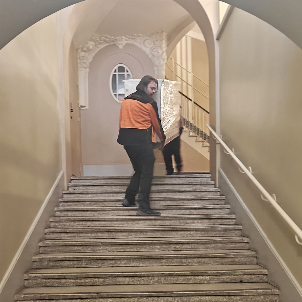 Muuttopalvelu Helsinki. Kaksi muuttomiestä kantamassa painavaa huonekalua rappukäytävässä Helsingin keskustassa.